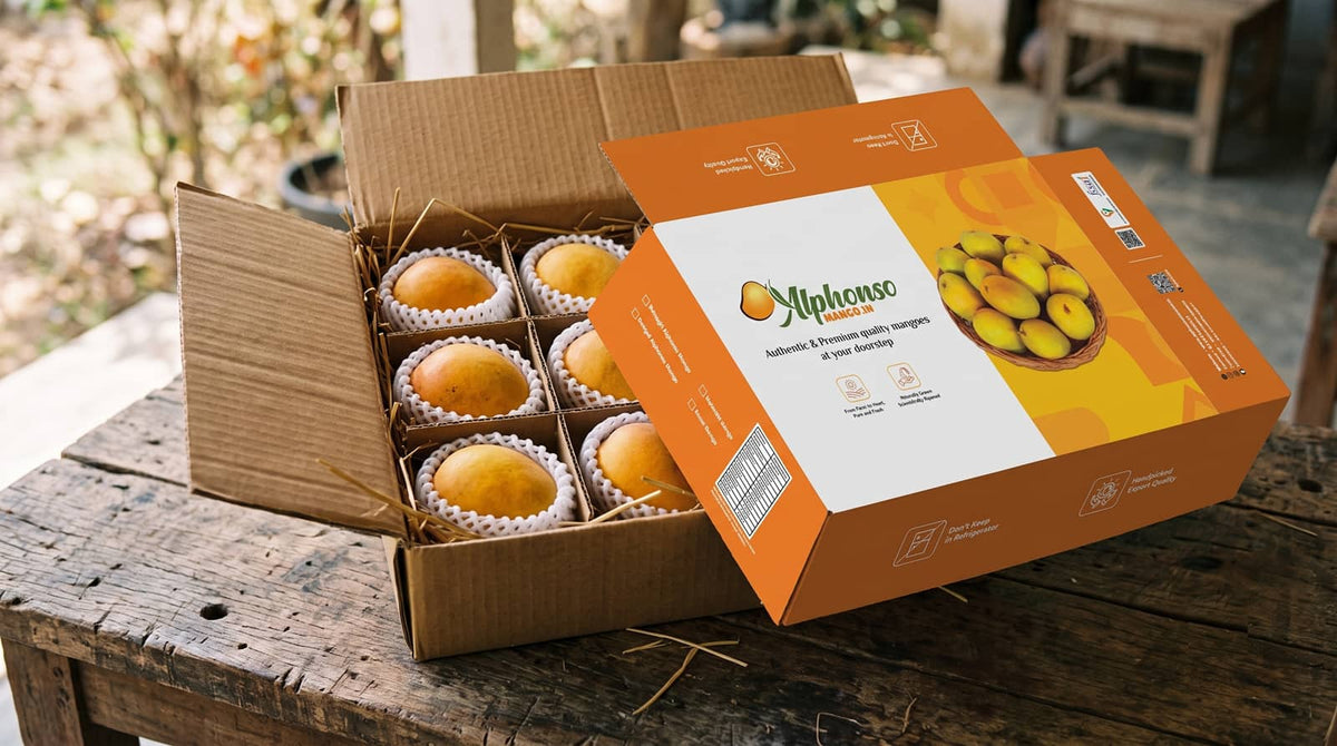 An open premium Alphonso mango peti packed with 12 ripe mangoes on straw, next to its branded cardboard top lid on a rustic wooden table.